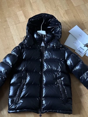 Mörkblå Moncler Maya med huva - Säljer en svart, glansig pufferjacka från Moncler med huva och två sidofickor med dragkedja. Jackan har quiltad design och synlig logga på ärmen. Perfekt för kalla dagar och har en snygg, modern look med full zip framtill.