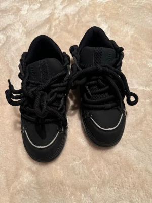 Svarta sneakers från Subshita - Chunky svarta sneakers från Subshita med grova snören och detaljer i silver. Skorna har en tjock sula i svart och grått, mesh och syntetmaterial samt rund tå. Märkeslogga bak på hälen och coola metalldetaljer framtill. Använda 1-2 gånger