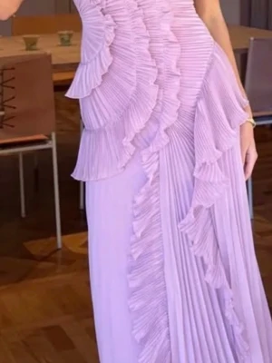 Ljuslila långklänning med volanger - Säljer en elegant ljuslila långklänning med plisserade volangdetaljer som går diagonalt över klänningen. Klänningen har en slits framtill och är ärmlös, vilket ger en snygg och modern look. Perfekt för dig som vill sticka ut med en unik design.