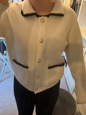 Vit kofta med svarta detaljer - Snygg vit kofta med svarta detaljer vid krage och fickor. Koftan har guldfärgade knappar framtill och två fickor med svart kant.