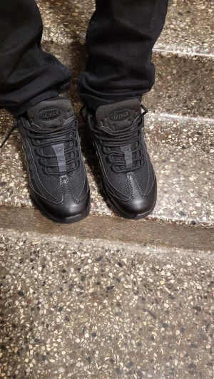 Nike Air Max 95 Svarta Sneakers - Nike Air Max 95 sneakers i helsvart färg med mesh och skinnpaneler. Snygg design med synlig Air Max-dämpning i sulan och rund tå. Skorna har snörning och en chunky platt sula som ger grym komfort. Perfekt för dig som gillar streetwear och sportig stil.