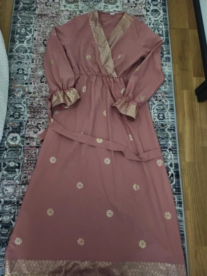 Rosa långklänning med blommigt mönster - Säljer en rosa långklänning med guldfärgade blommor och detaljer längs v-ringad. Klänningen har långa ärmar med volang och ett matchande bälte i midjan. 