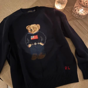 Polo Ralph L - Mörkblå stickad tröja från Polo Ralph L med ikonisk Polo Bear-motiv framtill. Tröjan har rund hals, långa ärmar och RL-broderi i rött vid nederkanten. Perfekt för dig som gillar klassisk stil med en twist. Tröjan är mindre i storleken så passar även M möjligtvis S