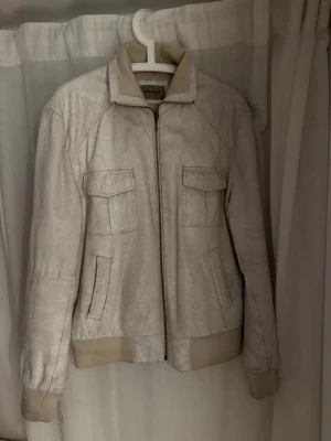 Beige bomberjacka skinn - Snygg beige bomberjacka i skinn med ribbade muddar och krage. Jackan har två fickor med lock framtill och två sidofickor, samt dragkedja hela vägen. Perfekt för dig som gillar stilren och enkel design.