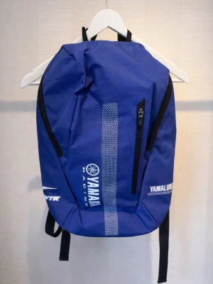 Blå Yamaha Racing ryggsäck - Säljer en blå Yamaha Racing ryggsäck med vita loggor och detaljer. Väskan har svarta dragkedjor, vadderade axelband och mesh på baksidan för extra komfort. sopass stor att du får plats med både laptop och träningskläder. Perfekt för dig som gillar motorsport och vill ha en snygg och praktisk väska.