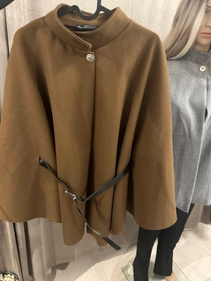 Brun cape-kappa med bälte - Snygg brun cape-kappa från Kuzary City med elegant silverfärgad knapp upptill och svart bälte i midjan. Kappan har en klassisk, vid form och hög krage som ger en stilren look. Perfekt för dig som vill sticka ut med en trendig och tidlös stil.
