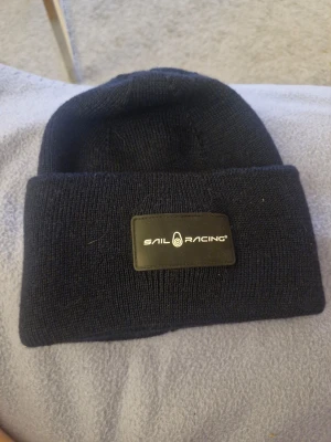 Mörkblå Sail Racing mössa - Snygg mörkblå mössa från Sail Racing med uppvikt kant och stilren logotyp framtill. Mössan är stickad och tillverkad i en blandning av ull och akryl, vilket gör den både varm och mjuk. Perfekt för kyliga dagar och enkel att matcha med din jacka.