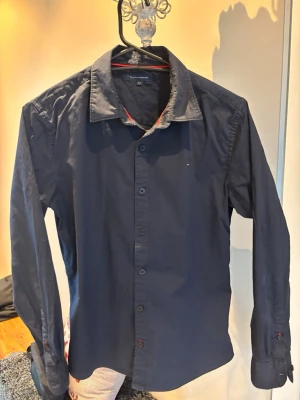 Mörkblå skjorta Tommy Hilfiger - Snygg mörkblå skjorta från Tommy Hilfiger med klassisk krage och diskret logga på bröstet. Skjortan har långa ärmar, svarta knappar och en röd detalj på insidan av kragen. Jag säljer denna då jag växt ut den men den är i ett otroligt bra skick. Kontakta mig vid intresse.