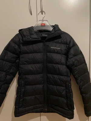 Svart Peak Performance dunjacka - Svart dunjacka från Peak Performance med huva och dragkedja framtill. Jackan har ett stilrent, quiltat mönster och logga på bröstet samt ärmen. Perfekt för kalla dagar och har en snygg, normal passform som funkar till många olika stilar.