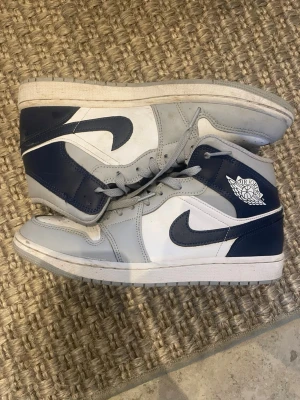 Nike Air Jordan 1 High vit och marinblå - Säljer ett par Nike Air Jordan 1 High sneakers i vit, grå och marinblå färg. Skorna har klassisk snörning, rund tå och platt sula. Ikonisk Air Jordan-logga på sidan och Swoosh i marinblått. Tillverkade i skinn med snygga paneler och kontrasterande färger.