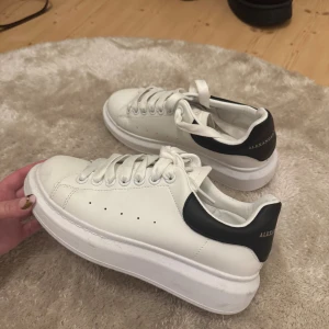 Alexander McQueen vita sneakers - Säljer ett par snygga vita sneakers från Alexander McQueen med svart häl och guldfärgad logga. Copi@. Skorna har bred plattformssula, rund tå och klassisk snörning. Använda fåtal gånger.