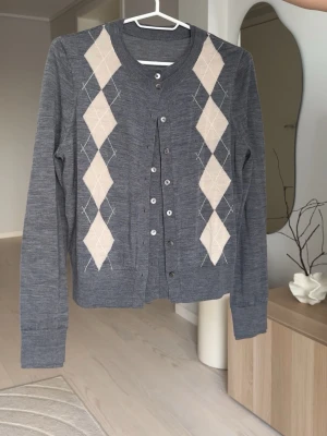 Grå kofta med beige argylemönster - Snygg grå kofta med klassiskt beige argylemönster framtill. Rund hals, långa ärmar och knappar hela vägen. Perfekt att styla över skjorta eller t-shirt för en preppy look. Mjuk och tunn stickad kvalitet som känns skön mot huden.