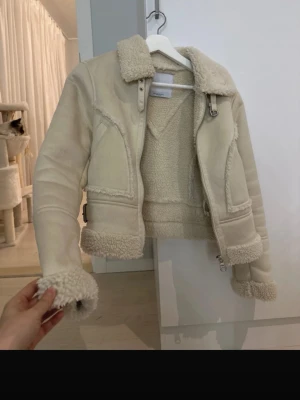 Beige teddyjacka från Bershka XS - Säljer en beige teddyjacka från Bershka i storlek XS. Jackan har fluffigt foder, detaljer med päls vid krage, ärmslut och nederkant samt dragkedja och spänne vid halsen. Perfekt för dig som gillar mysiga och trendiga jackor med coola detaljer.