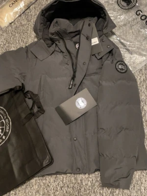 Mörkgrå dunjacka från Canada Goose - Säljer en mörkgrå vinterjacka från Canada Goose med huva och snygg svart logotyp på ärmen. Jackan har dragkedja och knappar framtill, samt flera praktiska fickor. Perfekt för kalla dagar och har en stilren look som funkar till många outfits. Storlek M\L