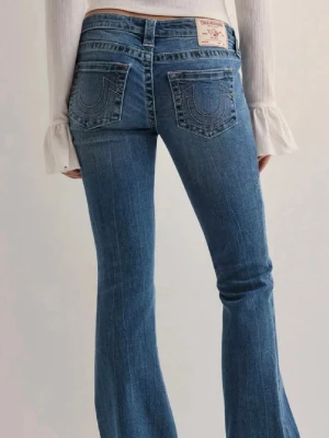 True Religion blå bootcut jeans - Snygga blå jeans från True Religion med klassiska broderade bakfickor och bootcut-snitt. Jeansen har låg midja, fem fickor och en patch med logga bak. Materialet är slitstarkt denim i bomull. Perfekta för dig som gillar en avslappnad och trendig look. Storlek W27!