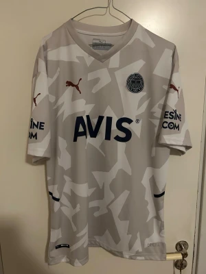 Fenerbahce tredjeställ 2021/22-puma-äkta  - Säljer en äkta Fenerbahce tredjetröja från säsongen 2021/22, tillverkad av Puma. Tröjan har ett stilrent beige/grått picelmönster, AVIS-logga på framsidan och Halley-tryck på ryggen. Tillverkad i lätt och bekvämt material med Puma DryCell-teknologi 