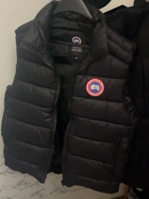 Svart dunväst från Canada Goose - Säljer en svart dunväst från Canada Goose med klassisk logga på bröstet. Västen har quiltad design, hög krage och dragkedja framtill. Perfekt för lager-på-lager och riktigt snygg till höst- och vinterlooken.