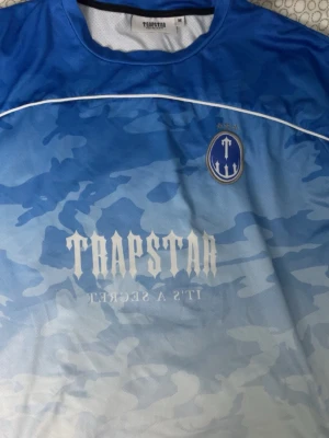  Trapstar t-shirt blå - Säljer en blå och ljusblå t-shirt från Trapstar med camomönster, stort tryck med siffran 23 på ryggen och Trapstar-logga både fram och bak. T-shirten har rund hals och är tillverkad i ett sportigt, ventilerande material. 