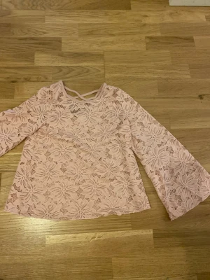 Rosa spetsblus från Lindex  - Säljer en ljusrosa blus från Lindex Kids i storlek 134/140. Blusen är helt i spets med stora blommönster, har långa utsvängda ärmar och en snygg v-ringning med korsade band framtill.