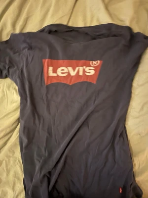 Svart Levi's t-shirt med röd logga - Säljer en svart t-shirt från Levi's med den klassiska röda loggan tryckt på bröstet. T-shirten har korta ärmar och rund hals. Materialet är mjuk bomull som är skön mot huden. Perfekt för dig som gillar streetwear och vill ha en ikonisk look.