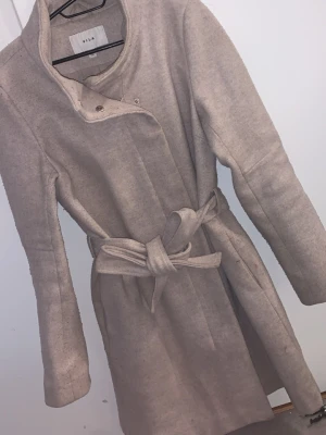Beige kappa med bälte från VILA - Snygg beige kappa från VILA med knytbälte i midjan och stilren krage. Jackan har långa ärmar och stängs med knappar upptill. Perfekt för dig som gillar en clean och klassisk look.
