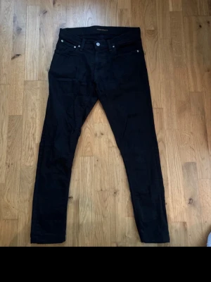 Svarta Nudie Jeans, storlek 31/32 - Säljer ett par svarta jeans från Nudie Jeans i storlek 31/32. Modellen har klassisk femficksdesign, svarta sömmar och Nudie-logga på bakfickan. Jeansen har en slim passform och är tillverkade i mjukt bomullsmaterial med lite stretch för extra komfort. Finns ett litet håll vid skrevet men är lätt att fixa