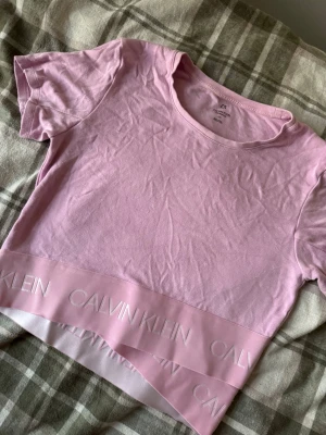 Ljusrosa topp från Calvin Klein - Säljer en ljusrosa topp från Calvin Klein Performance med korta ärmar och rund hals. Toppen har ett brett band längst ner med Calvin Klein-logga i relief. Materialet är mjuk bomull och modellen är croppad med normal passform.