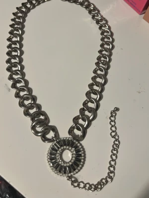 Silverfärgad chunky halskedja med rund hängare - Säljer en riktigt chunky halskedja i silverfärg med en stor rund hängare dekorerad med små vita och mörka stenar. Kedjan har breda länkar och justerbar längd med extra kedja. Perfekt för dig som gillar statement-smycken och vill sticka ut.