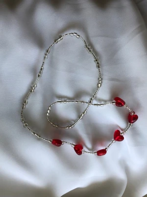 Handgjort halsband röda hjärtan - Unikt handgjort pärlhalsband med transparenta glaspärlor och röda hjärtformade plastpärlor som ger en gullig och lekfull design. Töjbar utan stängning. Perfekt accessoar för att lyfta din outfit med lite färg och kärlek. 