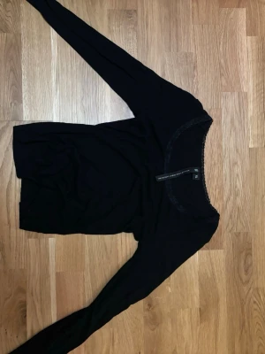 Svart långärmad topp med spetskant - Säljer en svart långärmad topp från The Black Label Collection i storlek XS. Toppen har en rund halsringning med fin spetskant som ger en extra detalj. Enkel och stilren design, perfekt att matcha med olika outfits.