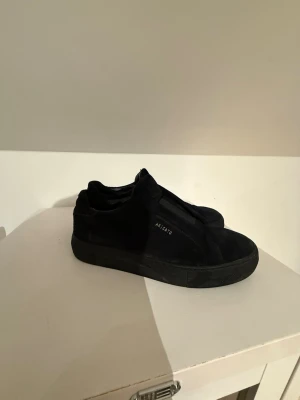 Svarta sneakers från Axel Arigato - Säljer ett par svarta sneakers från Axel Arigato med stilren design och diskret logga på sidan. Skorna har rund tå, platt sula och är tillverkade i mocka med elastiska paneler för enkel påtagning. Perfekta för dig som gillar minimalistisk stil. Lite stora i storleken, känns som de växer med fötterna 