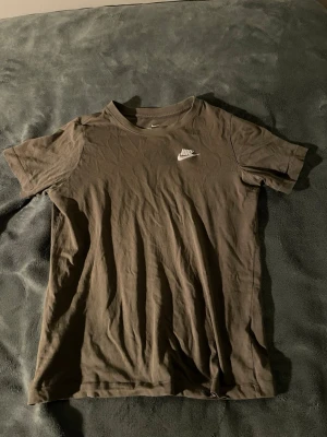 Brun grön Nike t-shirt i bomull - Säljer en mörkbrun t-shirt från Nike i storlek 147-158 . T-shirten har klassisk rund hals och korta ärmar, med en vit Nike-logga tryckt på bröstet. Materialet är mjuk bomull som känns skön mot huden. Perfekt för dig som gillar enkel och sportig stil.