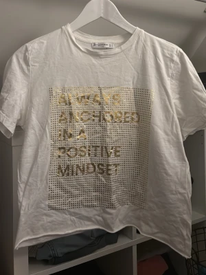 Vit t-shirt med guldtryck från Stradivarius - Snygg vit t-shirt från Stradivarius med korta ärmar och ett stort guldtryck framtill med texten 'Always anchored in a positive mindset'. Perfekt för dig som gillar statement-plagg och metallic-detaljer.