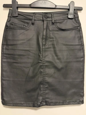 Svart skinnkjol från Noisy May - Säljer en svart kort skinnkjol från Noisy May med klassisk femficksdesign, bälteshällor och knappstängning fram.