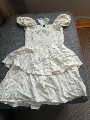 Vit blommig kortklänning H&M - Supersöt vit kortklänning från H&M med små gula blommor, puffärmar och smockad överdel. Klänningen har volangdetaljer. Perfekt för sommarens alla tillfällen! Passar även folk med S, säljer helt oanvänd och med prislappen kvar 