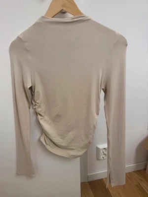 Beige långärmad topp från Gina Tricot - Säljer en beige långärmad topp från Gina Tricot i storlek S. Toppen har en fin passform och har snygga rynkade detaljer på sidan. Beige långärmad tröja i storlek S Polo krage  Inga skavanker Oanvänd