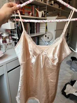 Champagnefärgad linnetopp med spets - Säljer en champagnefärgad linnetopp i glansigt satinliknande material med tunna axelband och spetsdetaljer vid urringningen och nederkanten. Toppen har en liten rosett framtill och är supermjuk och skön. Ett superfint och bekvämt pyjamas sett med topp och shorts