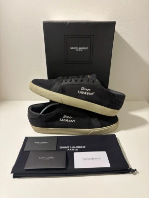 Saint Laurent skor - Säljer ett par svarta sneakers från Saint Laurent med vit logotyp på sidan. Skorna har snörning, rund tå och platt sula. Materialet är canvas med detaljer i skinn och en beige yttersula. Medföljer originalkartong, dustbag och äkthetskort. Priset är inte hugget i sten!