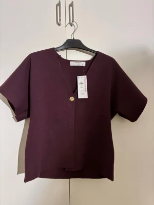 Vinröd blus från Primark - Säljer en stilren blus från Primark i vinröd färg. Blusen har korta ärmar, v-ringad omlottfront med en dekorativ guldfärgad knapp. Materialet är mjukt och har en lätt struktur.