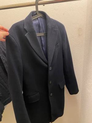 Mörkblå ullkappa från Mango Man - Snygg mörkblå kappa från Mango Man i klassisk modell med tre knappar framtill och två fickor. Jackan är helfodrad och har en stilren krage. Perfekt för dig som vill ha en tidlös och clean look.