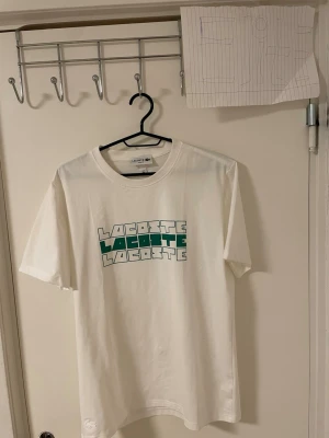Vit Lacoste t-shirt med tryck - Vit t-shirt från Lacoste med coolt grafiskt tryck i grönt och vitt på bröstet. Klassisk rund hals och korta ärmar. Materialet är mjuk bomull som känns skönt mot huden. Perfekt för dig som gillar stilrena och sportiga vibes.Liknande modell som originala.