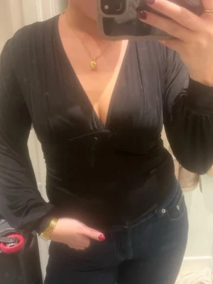 Svart omlottopp med v-ringning - Säljer en svart omlottopp med djup v-ringning och långa ärmar. Toppen har en figurnära passform och är tillverkad i ett mjukt, glansigt material som ger en elegant känsla. Perfekt att styla med jeans eller kjol för en snygg look.