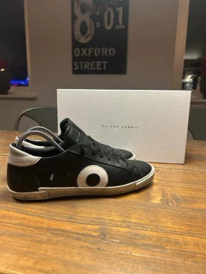 Svarta sneakers från Oliver Cabell - Säljer ett par svarta sneakers från Oliver Cabell med vit detalj och rund cirkel på sidan. Skorna har snörning, vit kant vid hälen och platt sula. Materialet är skinn och de har en klassisk, rund tå. Perfekt för dig som gillar stilrena och coola sneakers.