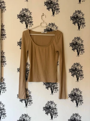 Beige ribbad långärmad topp NA-KD - Säljer en beige långärmad topp från NA-KD med ribbad struktur och rundad, djup urringning. Modellen är figurnära och har en enkel, stilren design som funkar till många olika looks. Perfekt att styla med jeans eller kjol.
