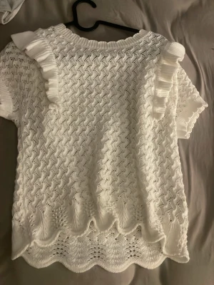 Vit stickad blus med volang från Lindex - Supersöt vit stickad blus från Lindex med korta ärmar och volangdetaljer på axlarna. Blusen har ett vågigt mönster och ribbad halskant, perfekt för dig som gillar romantisk och feminin stil. Passar dig som vill ha något extra till vår och sommar.