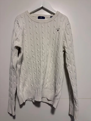 Vit kabelstickad tröja från GANT - Säljer en vit kabelstickad tröja från GANT med broderad logga på bröstet. Tröjan har rund hals, långa ärmar och ribbade muddar vid ärmslut och nederkant. OBS! Liten i storleken (snarare som 140)