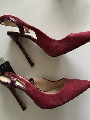 Vinröda pumps med ormskinnsdetalj från Zara - Säljer ett par vinröda pumps från Zara i äkta skinn med snygga ormskinnsdetaljer på klack och rem. Skorna har spetsig tå och hög klack, vilket ger en riktigt elegant vibe. Materialet är mocka och skinn, och färgen är djup vinröd med mörka toner. HELT NYA med etikett. Nypris 1299:-. 