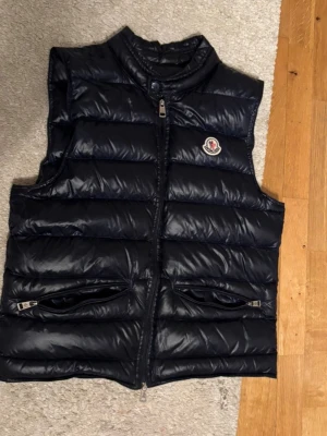Mörkblå  dunväst från Moncler - Säljer en svart dunväst från Moncler med klassisk logga på bröstet. Västen har quiltad design, två fickor med dragkedja och hög krage. Materialet är glansig polyester och dunfyllning som håller värmen. Perfekt för lager-på-lager under kyliga dagar. Storlek 1