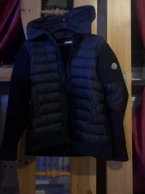 Svart Moncler pufferjacka med huva - Säljer en svart pufferjacka från Moncler med huva och dragkedja. Jackan har quiltade partier framtill och på ryggen, samt släta stickade ärmar med Moncler-logga på armen. Praktiska fickor med dragkedja och en clean, modern look. Nfc på armen fungerar utmärkt!