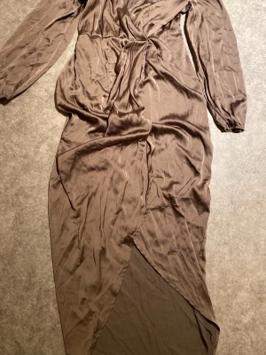 Brun omlottklänning med lång ärm - Säljer en brun långklänning med omlott-design och knyt i midjan. Klänningen har långa ärmar med resår vid handlederna och är gjord i ett glansigt, mjukt material som ger en lyxig känsla. Perfekt för dig som gillar stilrena och eleganta plagg.